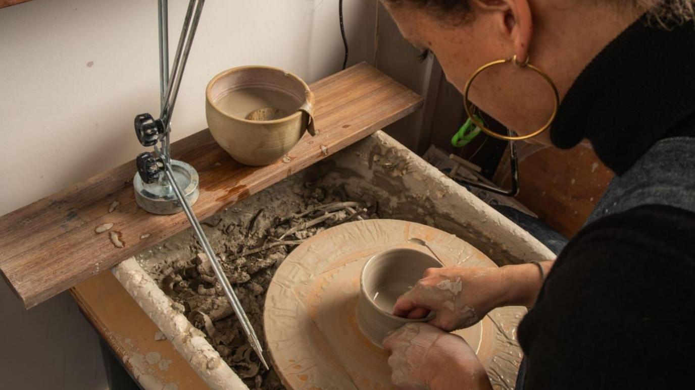 A woman in a black top and gold hoop earrings moulding a clay pot on a pottery wheel