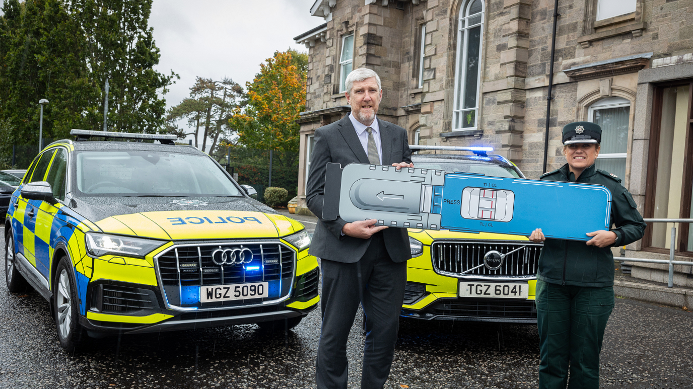 John O'Dowd in a black suit and a grey shirt holds a large drug testing kit alongside a female police officer, in uniform, in front of two police cars