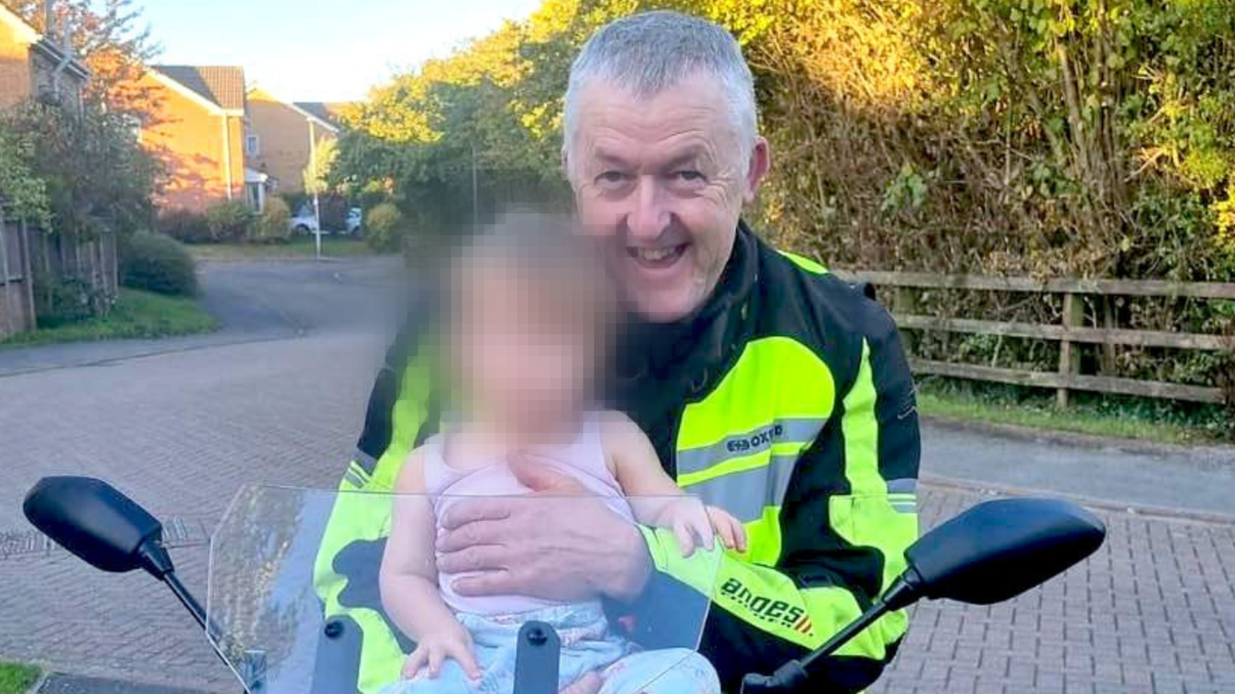 Motorcyclist, Nigel Osler, sat on a stationary motorbike in a driveway, carrying a baby whose face is blurred