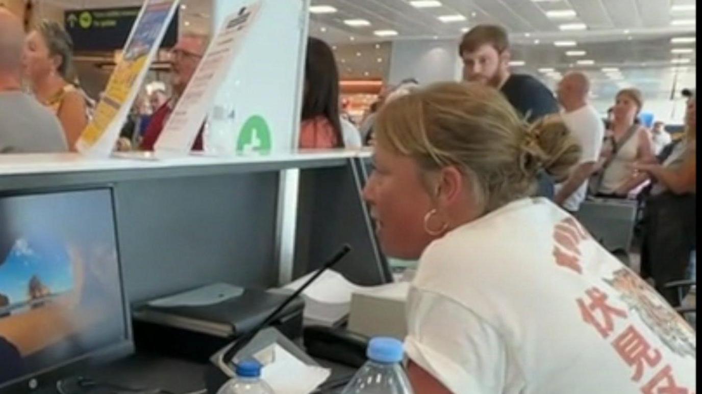 Faye Williams behind the departure desk speaking into the tannoy. She is wearing a white t-shirt. 