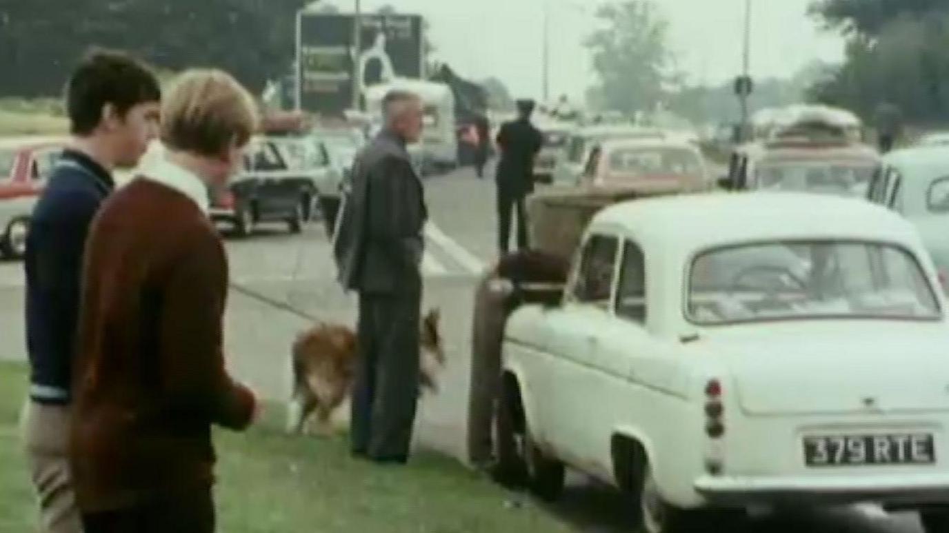 An archive image of a busy road with traffic at a standstill. The image was taken in 1974 and there are cars of that period queuing on a main road, with people waiting at the side of the road. One man in a grey suit has a dog on a lead. 