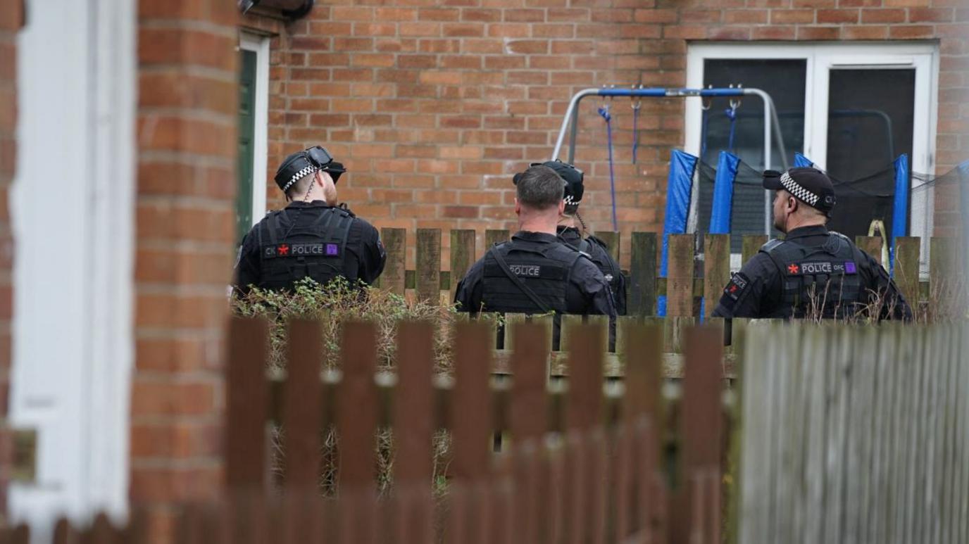 Four armed policemen in black uniform are seen in a garden with fences in the foreground and a swing in the background, surrounded by houses