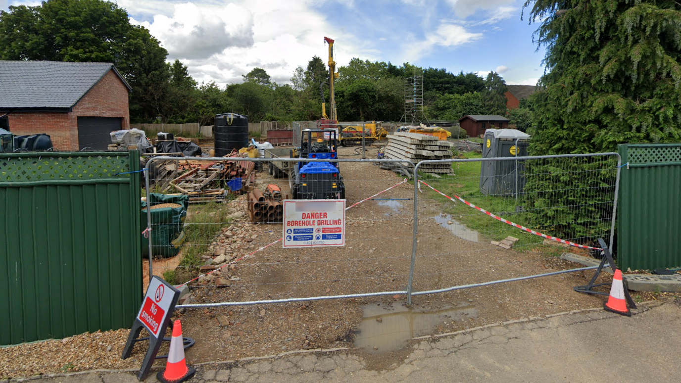 A driveway to a property that has been turned into a construction site, containing construction vehicles, metal pipes and a portable toilet. The sign on the metal fence that cordons off the site says, "Danger, borehole drilling".