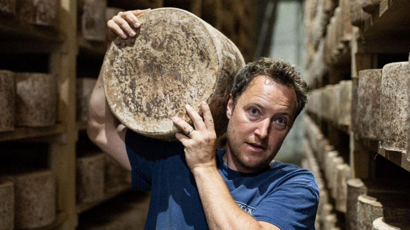 A man stands in a warehouse with shelves stacks with large wheels of a cheddar cheese, on of which he is holding on his shoulder.