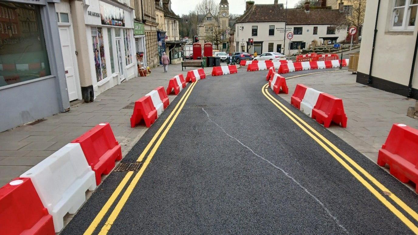 A newly-tarmacked road on a town's high street with freshly-painted double yellow lines and red and white barriers alongside it.