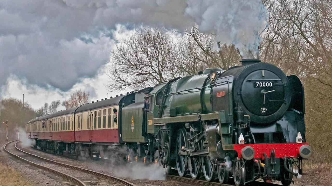 Britannia 70000, a green steam engine with plumes of smoke billowing from its funnel, pulling carriages on a railway track