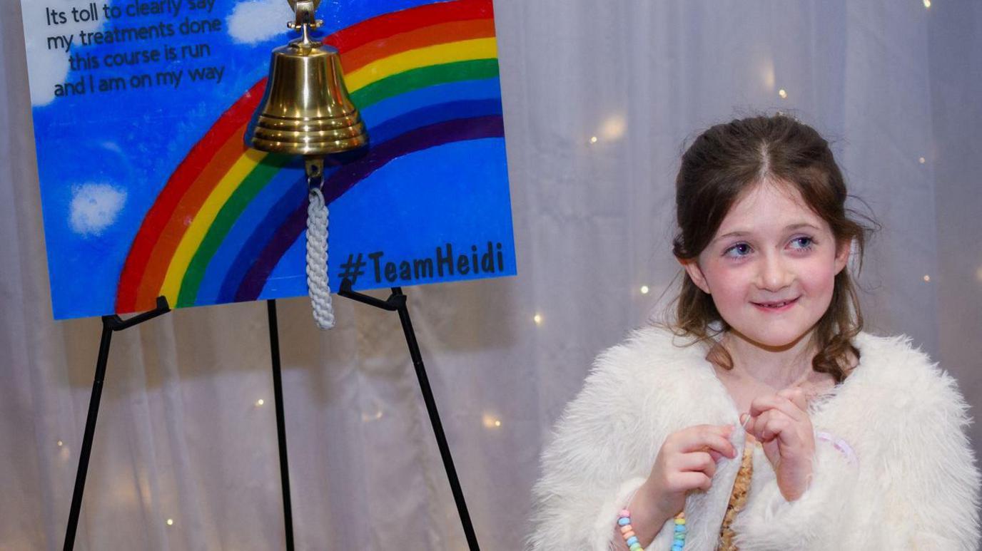 A young girl stands in front of a sign that reads "its toll to clearly say my treatments done this course is run and I am on my way". A bell hangs in front of it.