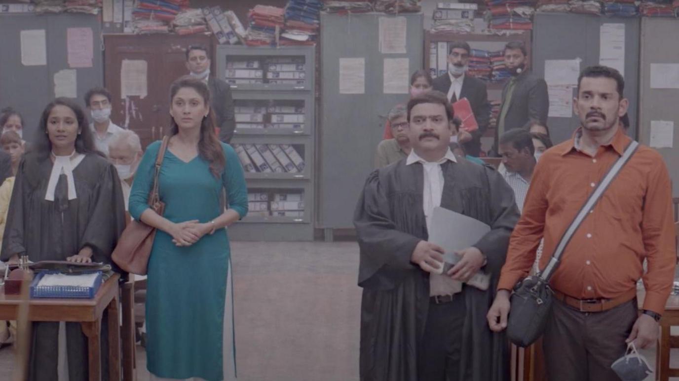 A still from the film showing a woman wearing a blue kurta standing with her lawyer in a courtroom. To her right, her husband stands wearing an orange shirt and crossbody bag, with his lawyer standing next to them. A thin layer of polluted air hangs in the room