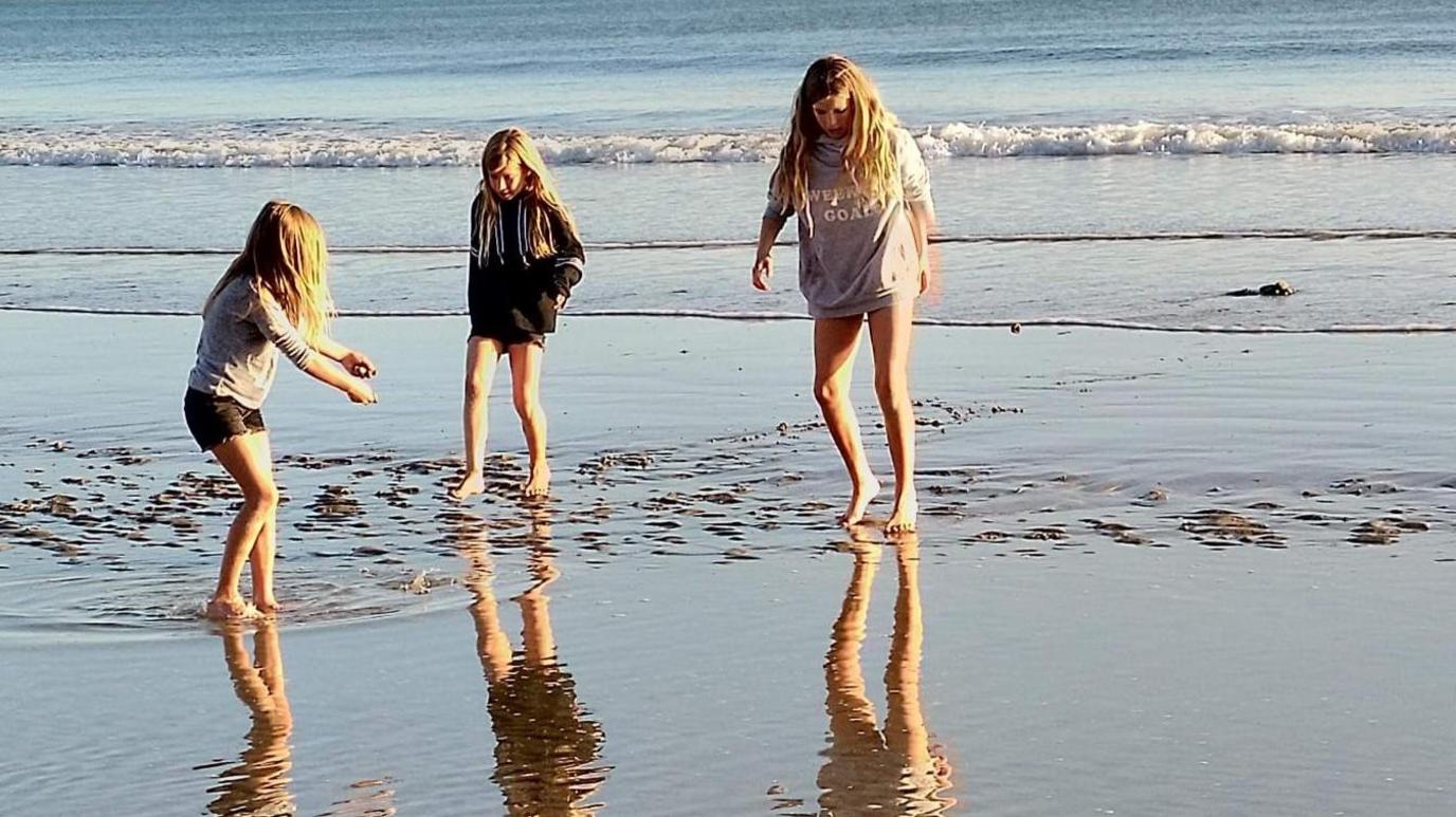 Three young girls wearing shorts and sweatshirts paddle in water on a shoreline. All three have long blonde hair