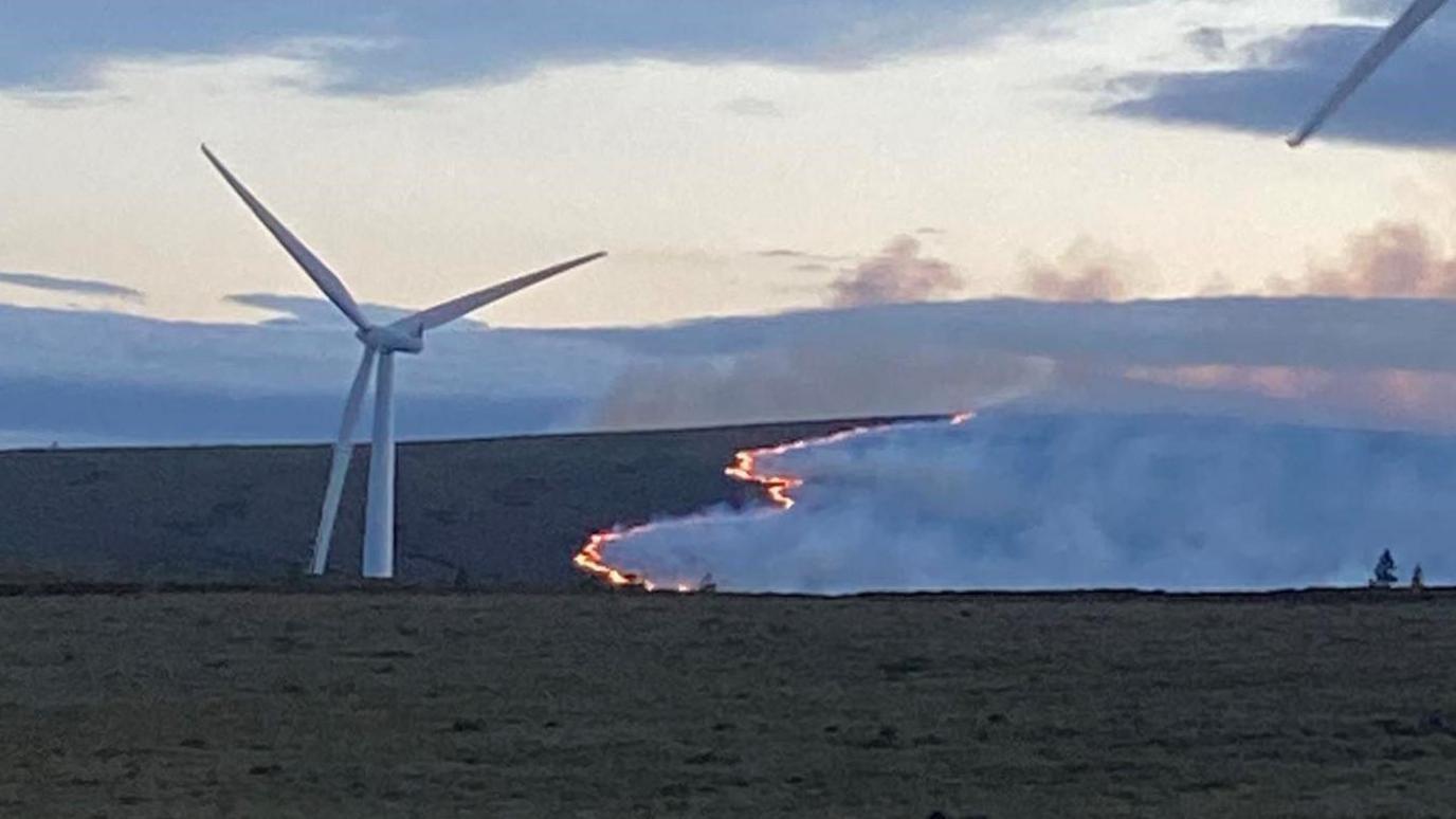 Smoke drifts from a line of flames on a large moorland. A wind turbine is in front of the flames.