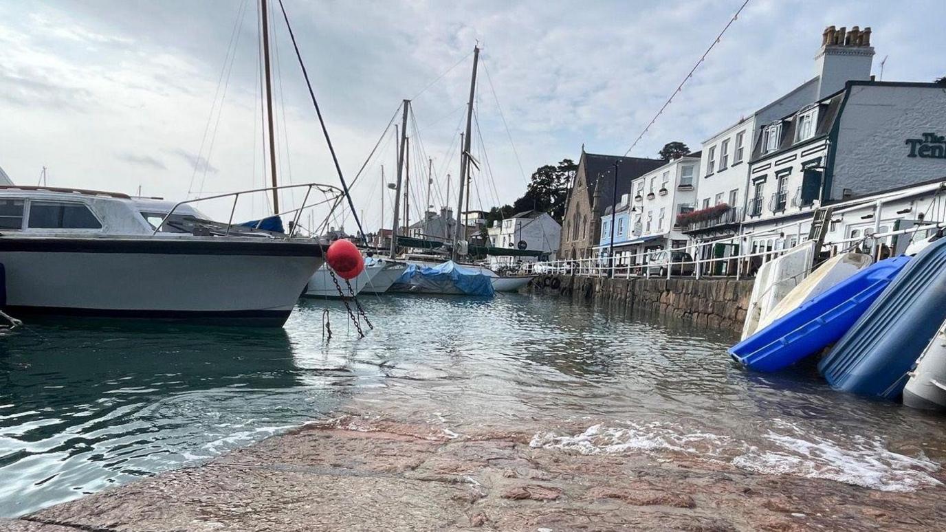 The walkway in St Aubin's harbour in St Brelade is covered with sea water. There are yachts moored and dinghies resting on a section of seawall with properties lining a street above. 