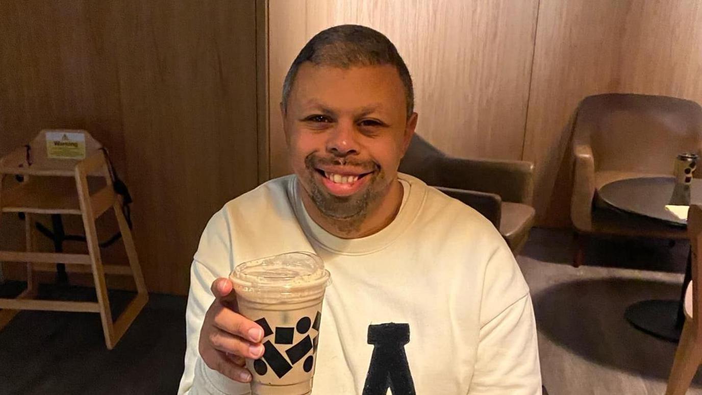 Reece Kinsman sitting in a cafe. He is wearing a white jumper, holding a coffee and is smiling. He has short hair and some facial hair.