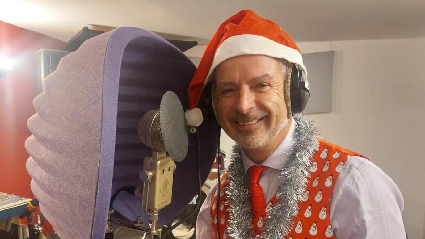 Simon Billings wearing a red and white santa hat, a red waistcoat with snowmen on it and a white shirt. He has silver tinsel wrapped around his neck and he is smiling. In front of him is a microphone and associated noise cancelling equipment.