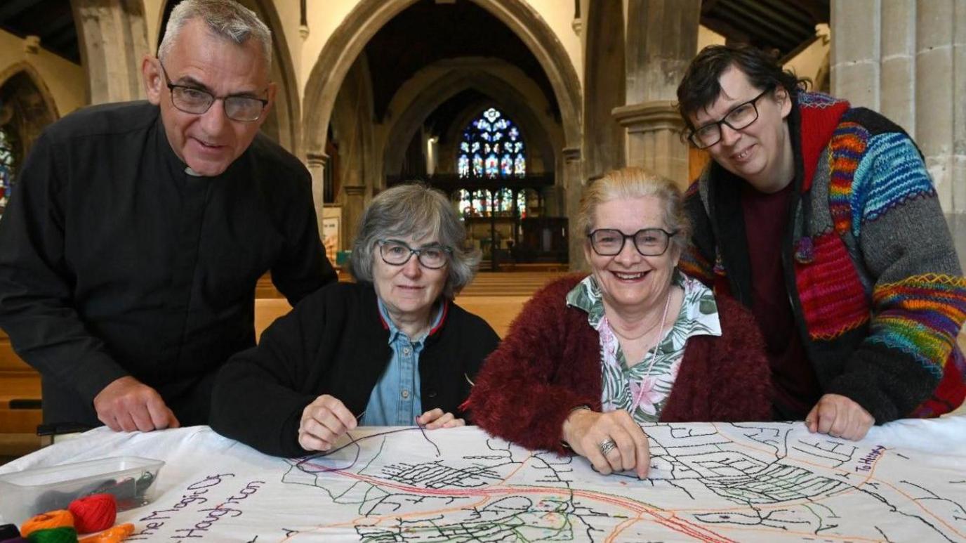 The Reverend John Barron (left) wearing a traditional black shirt and dog collar and the Reverend Claire Cullingworth (right) wearing a multi-coloured knitted chunky cardigan. They are pictured with volunteers Denise Hall and Debbie Anderson between them, alongside the Map of Houghton at St Michael's and All Angels Church. The map is stitched on to a white fabric sheet.
