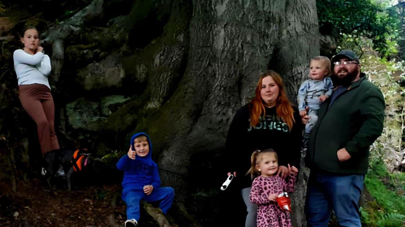 Nicole and Darren stand under a tree. Darren holds their youngest son, their youngest daughter stands in front of Nicole. One of their sons sits next to them on a tree branch, their eldest daughter leans on a large tree root at the edge of the photo. Their middle daughter stands on top of the tree roots