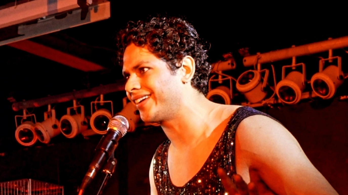 Vyome Singh is talking into microphone, which is being held up on a stand, and is standing in front of theatre lights, which are all turned off.