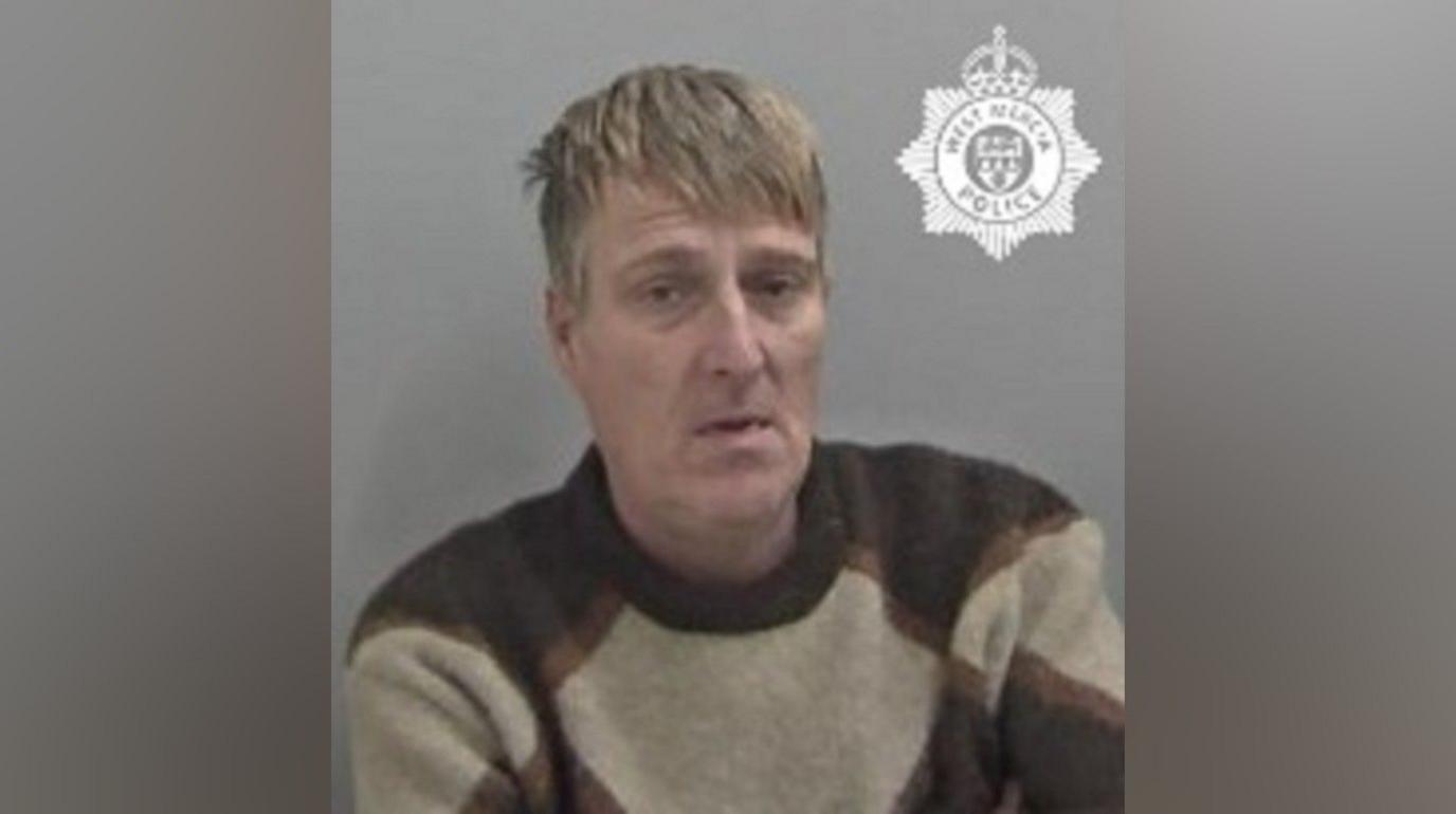 Craig Sawyer is seen in a police mugshot. He is wearing a beige and dark brown jumper and has dishevelled hair and frown lines. He is leaning slightly to one side.