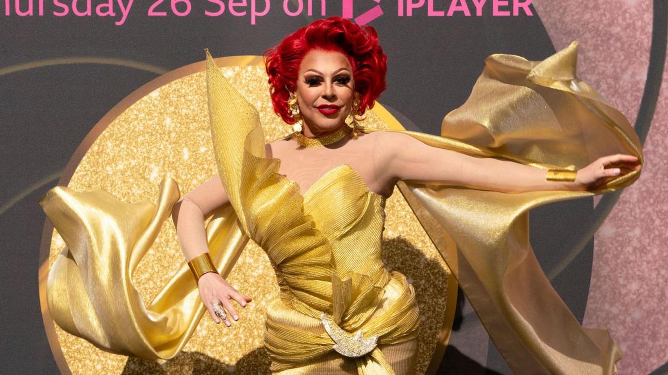 La Voix is wearing a large gold dress with a large glittery circular back at a launch event for RuPaul's Drag Race UK.