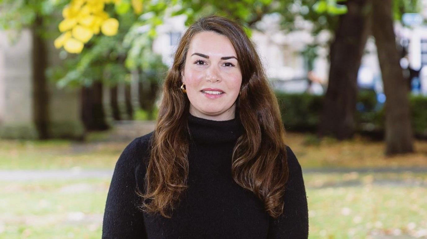 Stephanie Mansell-Jones is standing in front of grass and trees. She has long brown hair and is wearing a black jumper.