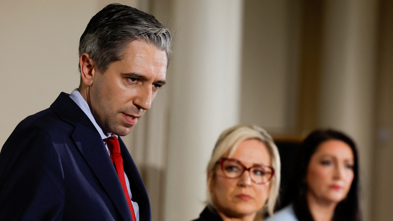 Simon Harris, wearing a suit, blue shirt and red tie, to the left of the photo. Michelle O'Neill and Emma Little-Pengelly are in the blurred background of the photo. 