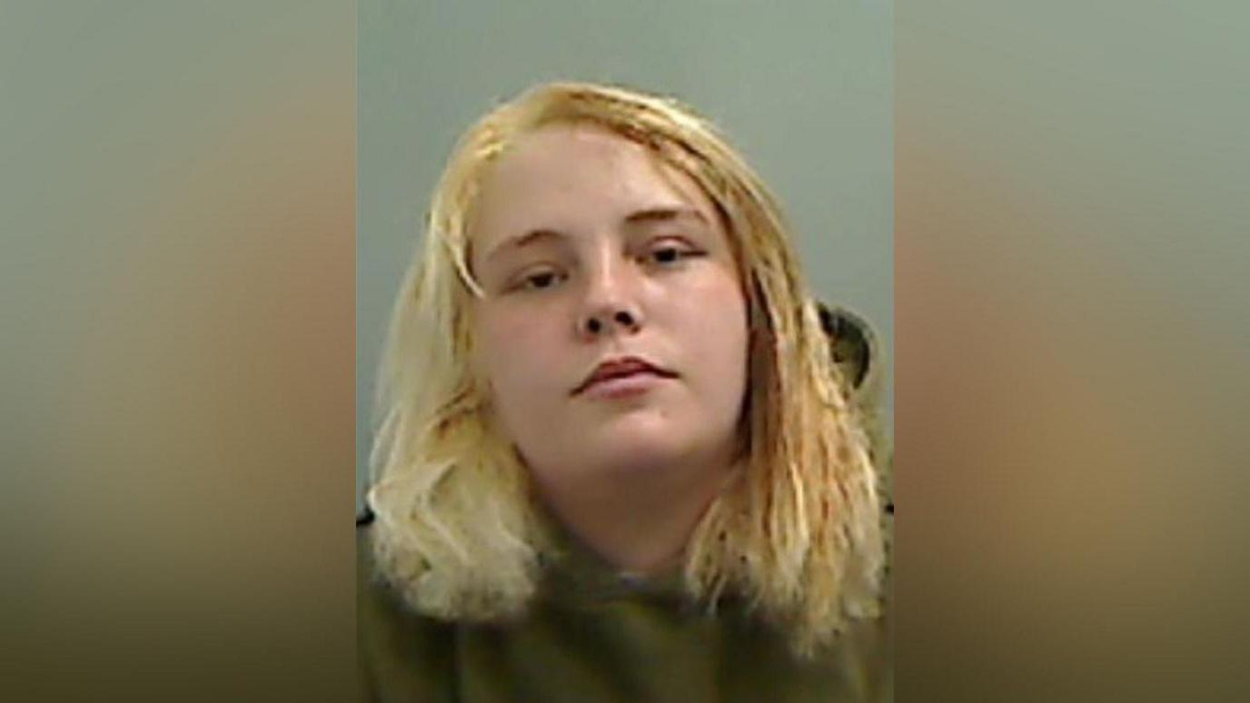 Mugshot of Cerr. She has blonde hair and is wearing a green hoody