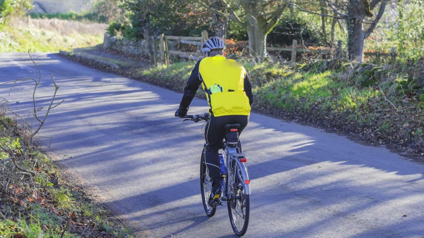 A man in a yellow hi-vis jacket cycling along a country lane.