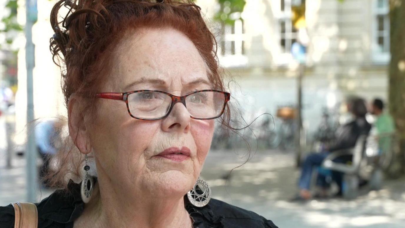 Caroline has red hair and is wearing glasses and silver earrings and a black top. She is standing in the street on a sunny day.