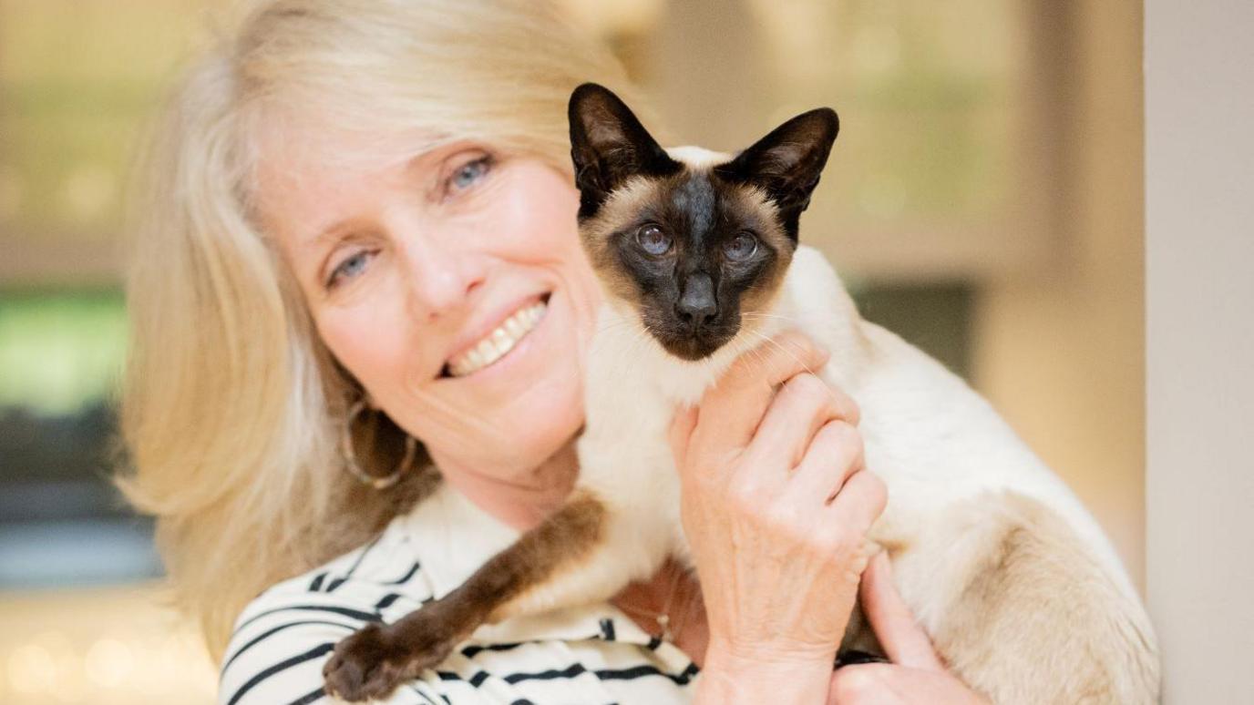 A woman in a striped shirt holding a Siamese cat indoors.