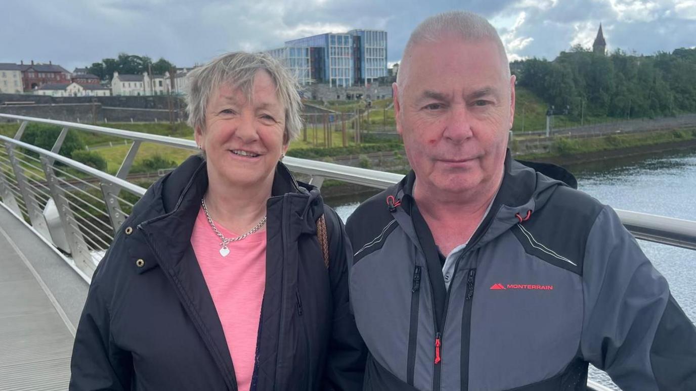a man and woman stand side by side on a bridge, a river is behind them, and a number of buildings can be seen in the background