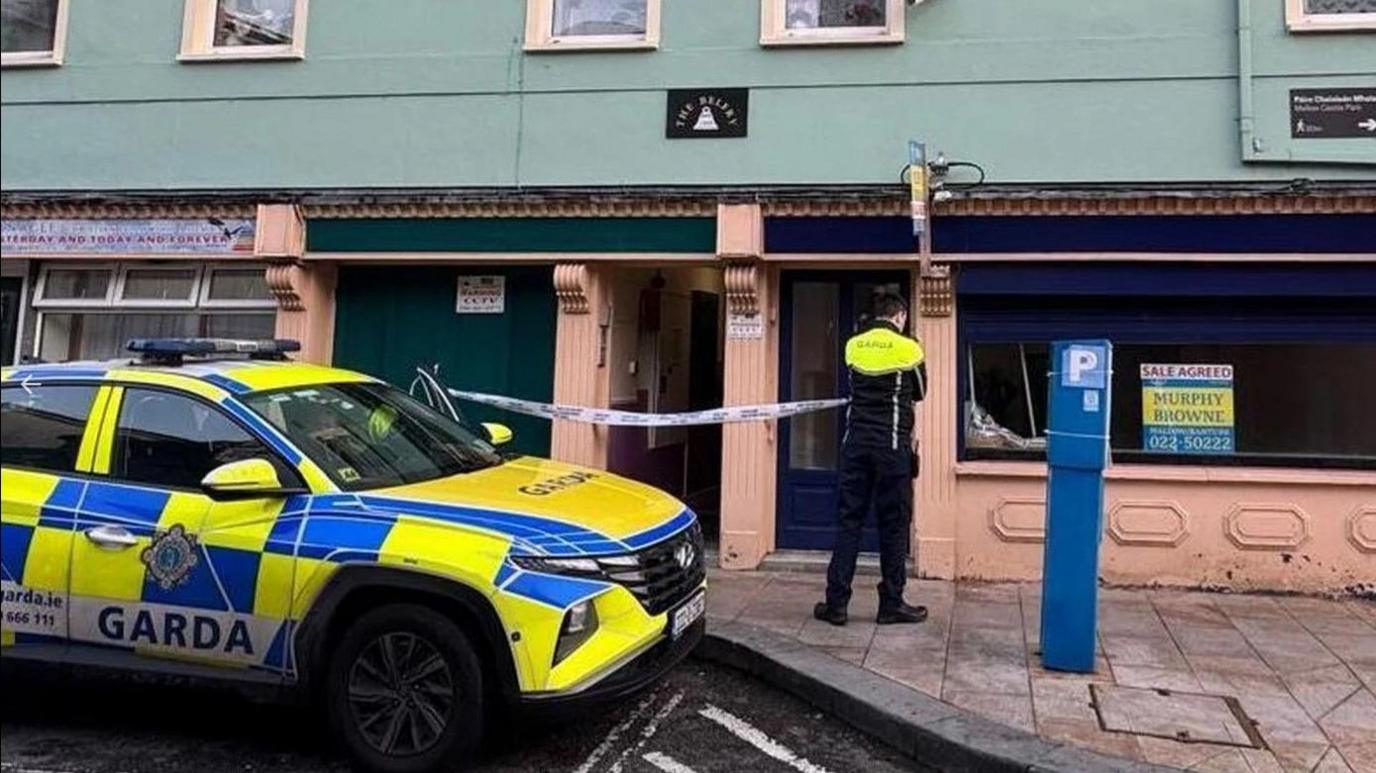 A yellow and blue Gardai car sits on the black tarmac outside a green and peach brick building. One officer with a Yellow and black uniform stands with his back to the camera, outside the premises.