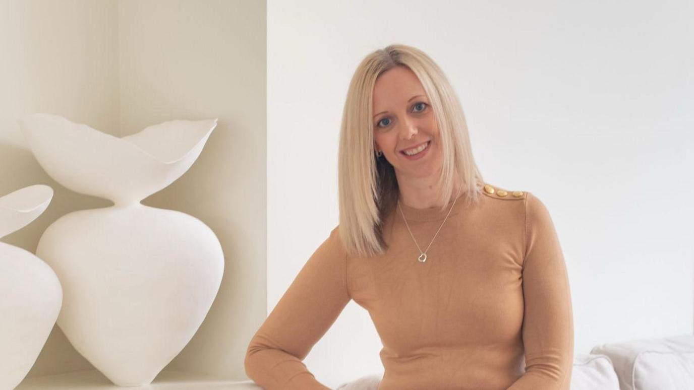 A blonde woman wearing a camel-coloured turtle neck t-shirt and smiling at the camera