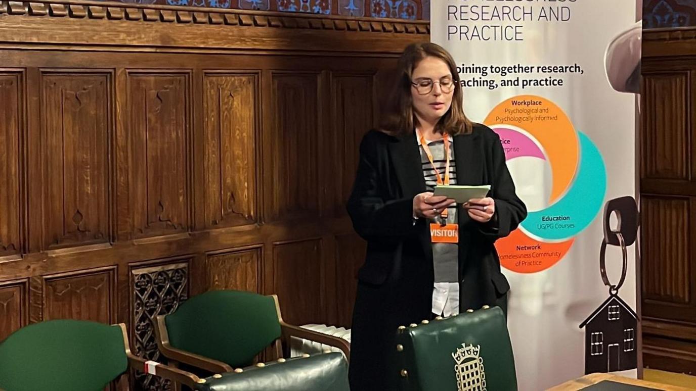 A young woman with brown hair, wearing a black coat and glasses. She is standing in a room in the Houses of Parliament, in front of a wood-panelled wall and a head-height poster featuring information from the Step by Step charity. She is wearing glasses and  reading from a notepad which she is holding in her hands in front of her. Beside her are two dark green chairs. She has an orange lanyard on which reads "visitor".