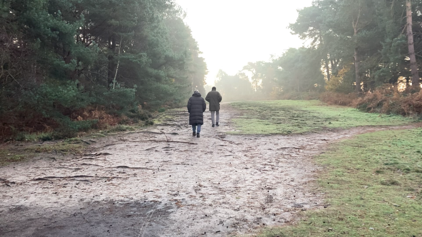 Two people walking in Sutton Heath woods away from the camera. They are wearing coats. There are trees lining the footpath either side. It looks like a frosty but sunny winter's day.