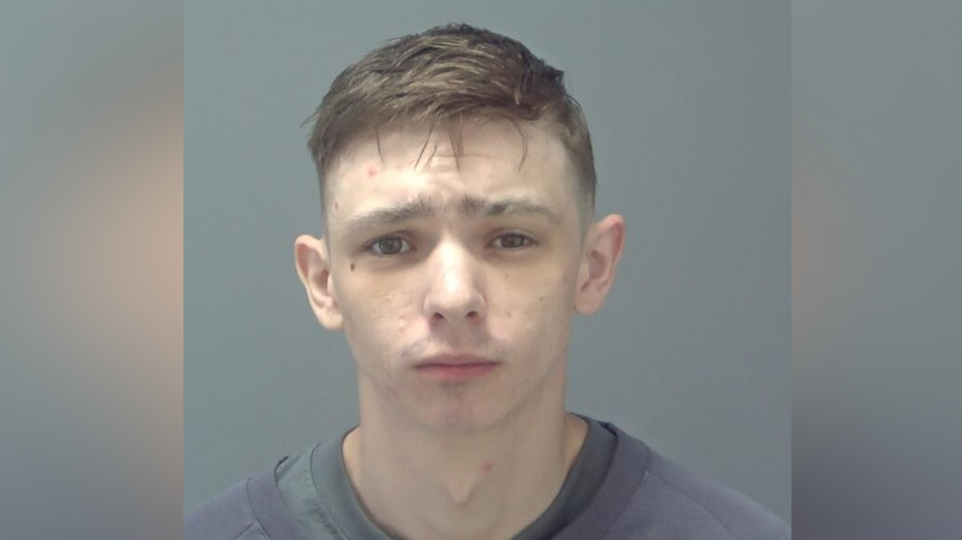 A police custody photo of a man with short brown hair. He is wearing a blue jumper with a grey top on underneath.