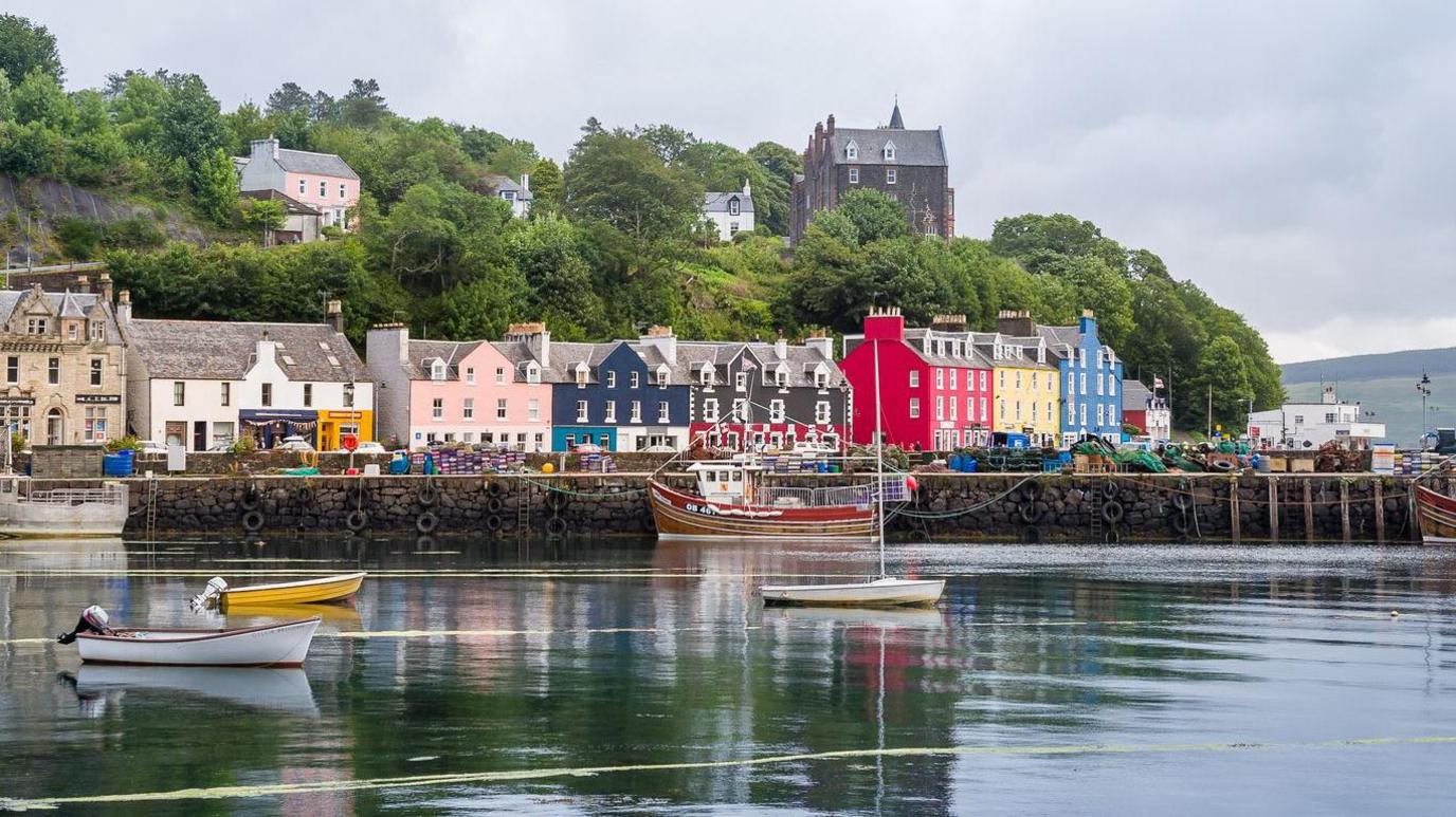 A view of the colourful buildings in Tobermory, Mull taken from across the harbour. There are small boats in the water and trees on a hill behind the buildings.