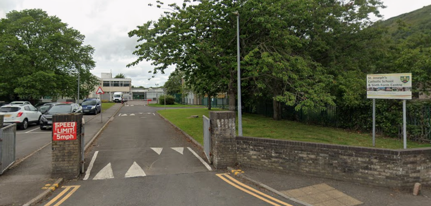 A general view from the road of St Joseph's Roman Catholic Comprehensive School in Aberavon. A gate is open leading into the school grounds with a waist-height stone wall surrounding the school boundary. A white sign with the school name on can be seen in the left hand side. Through the gate is a car park filled with cars with the school buildings in the background. 