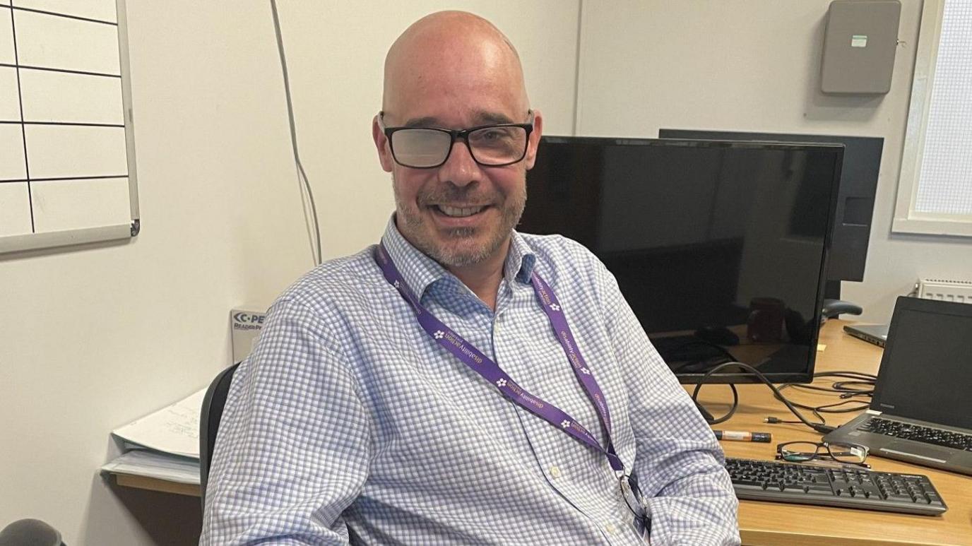 A bald man sitting in an office chair at a desk, he is wearing a blue and white checked shirt and has a purple lanyard around his neck. He is wearing a pair of black framed glasses, his right glasses' lens is clouded whilst the left lens is transparent. To the man's right is a support stick which he is not holding.