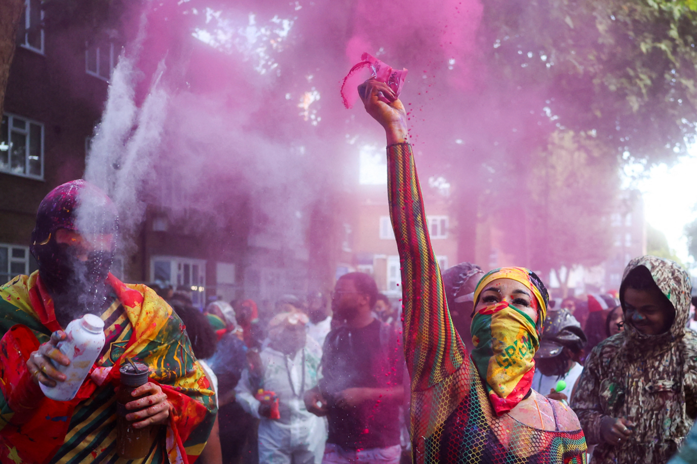 A woman throws pink paint powder into the air in the street with other revellers around her