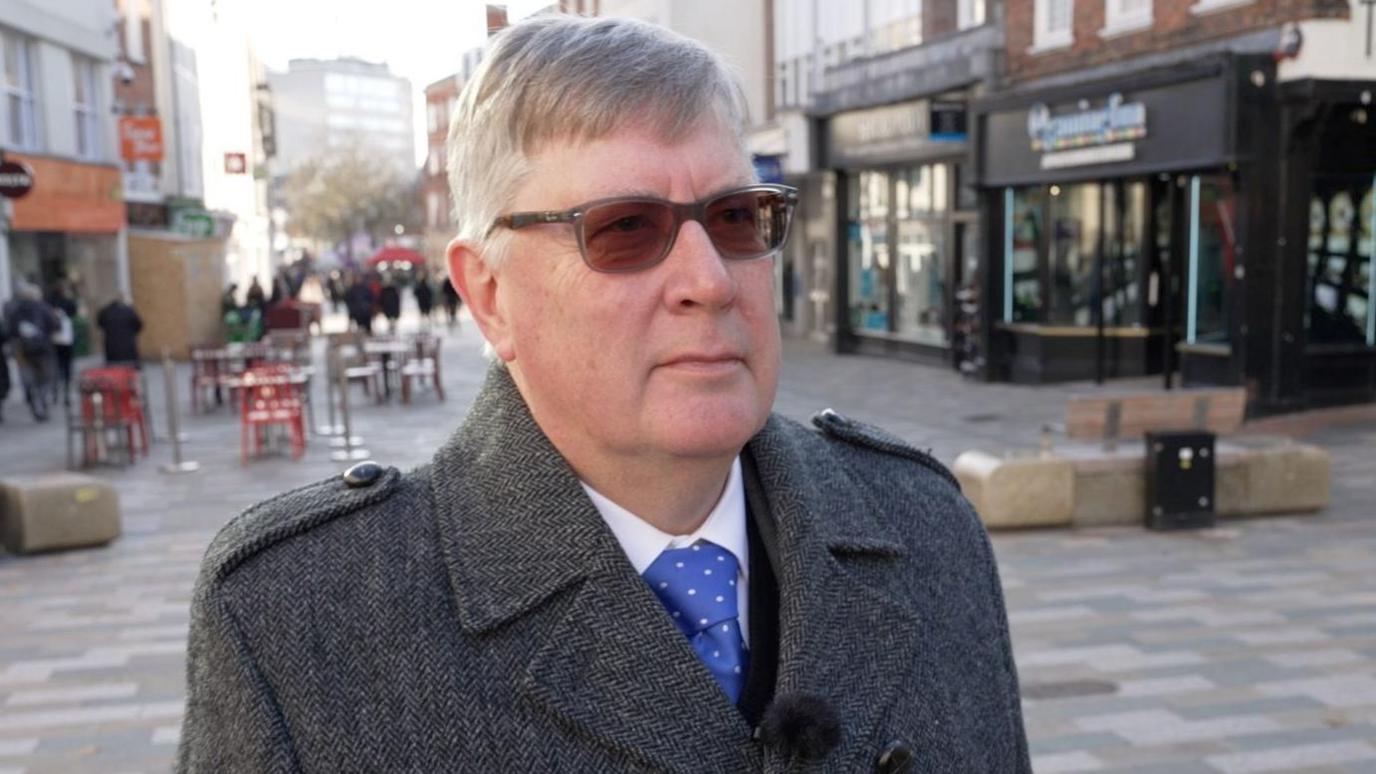 Roger Hirst is standing in Chelmsford high street with sunglasses on. He is looking slightly in front of the camera. He is wearing a grey jacket and a white shirt with a blue tie. He is not smiling. His hair is grey and combed to the right side of his face to give him a fringe.