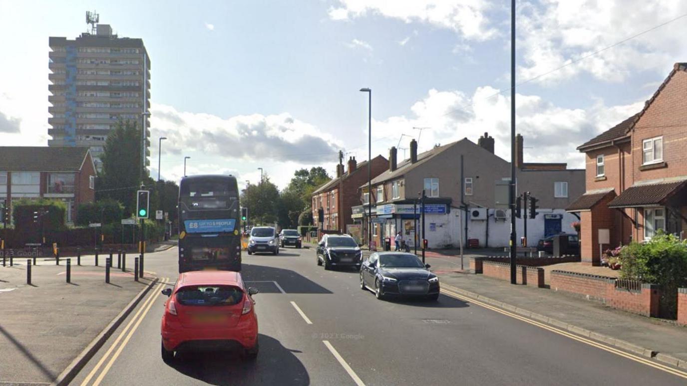 Bell Green Road in Coventry. It is an overcast day. The road has a number of cars on, including a bus. A corner shop and some houses can be seen. 
