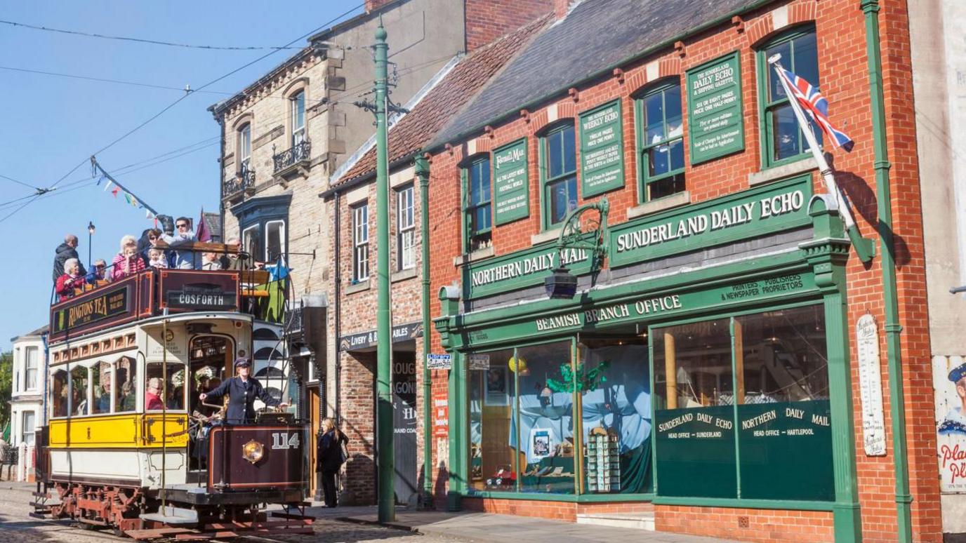 View of a tram dating from Edwardian times with a number of people on board and a conductor in front of the spiral steps leading to the top deck. It is in front of a shop from the same era, Beamish Branch Office, with double fronted plate glass windows. Boards above the windows advertise the Northern Daily Mail and Sunderland Daily Echo