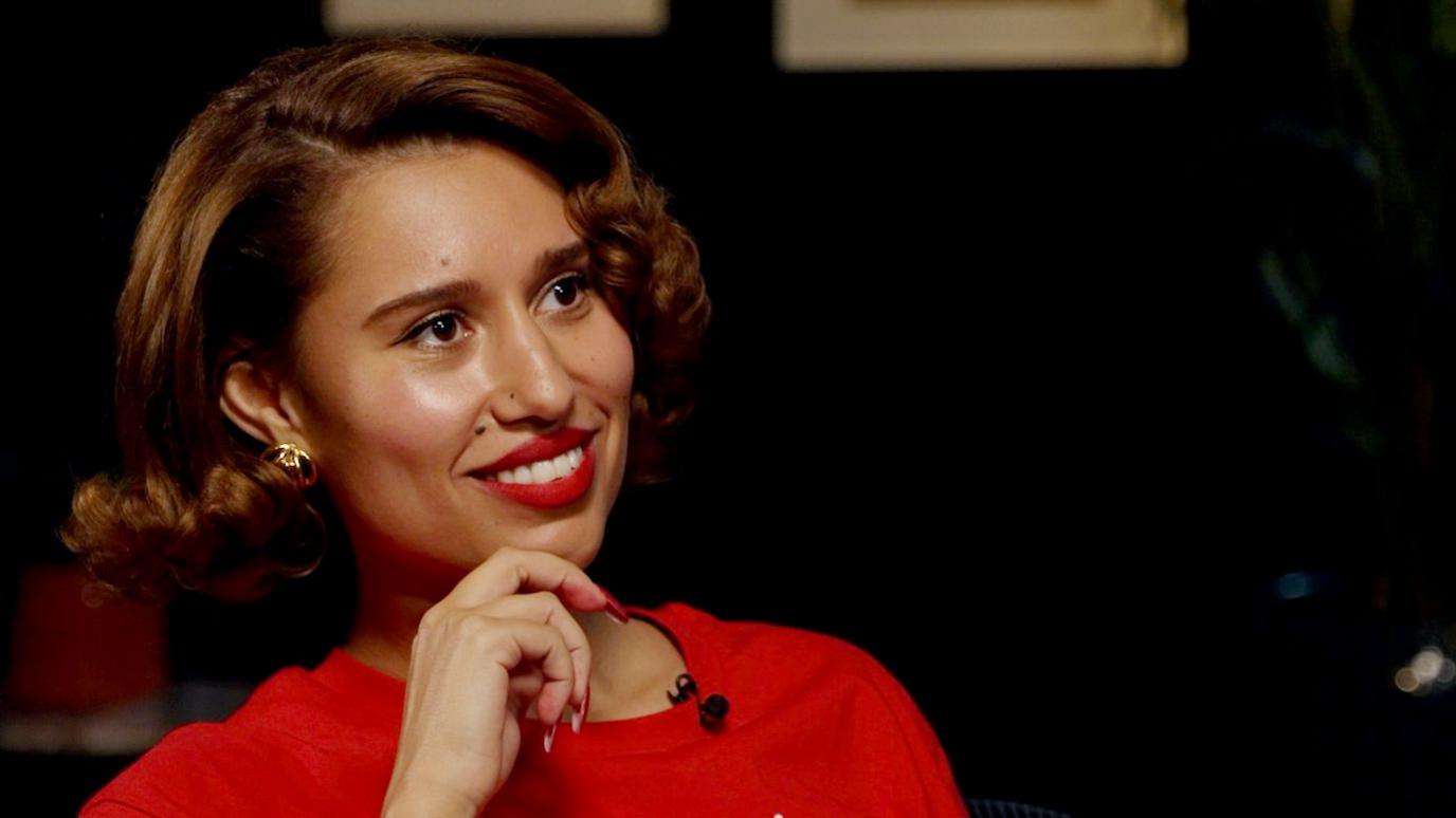 A close up head and shoulders crop of an image of Raye as she smiles and looks away from camera while touching her chin with her hand and wearing a red T shirt against a black background.