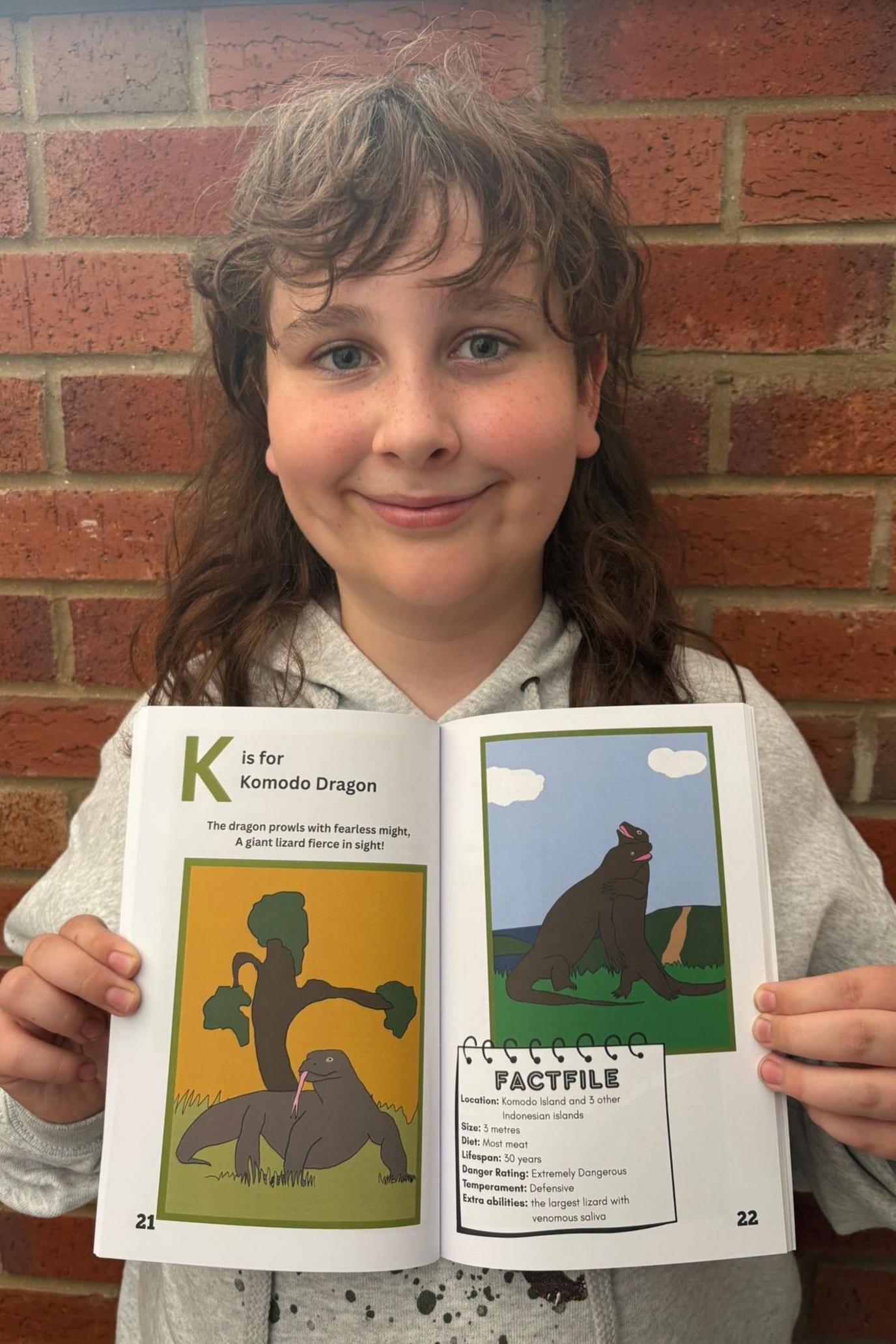 A young boy with long brown hair and a fringe is smiling into the camera and holding up an open book. The book says 'K is for Komodo Dragon' and has two colourful drawings and a fact file on the right. He is standing against a brick wall and wearing a grey hoodie with black splatters on it.