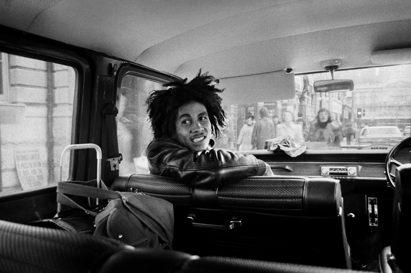 Black and white photograph of Bob Marley in the passenger seat of a van with his body partially turned towards the back of the vehicle