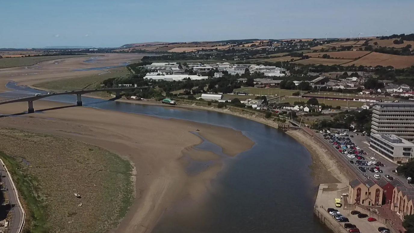 A drone shot of Barnstaple in Devon. It shows a big bridge crossing a river that is lacking water and mainly sand. There are houses and lots of cars parked along the quay side. There is also clear blue skies.