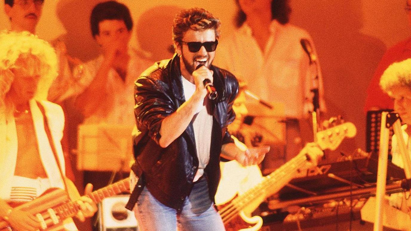 George Michael singing at Live Aid in a leather jacket and sunglasses, with the band behind him under amber stage lighting.