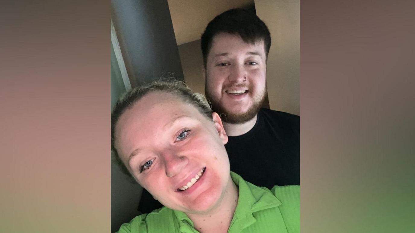 A selfie-style image of a woman, with blonde hair in a ponytail and a green top, who is standing in front of a man with dark hair and a black T-shirt. They are both smiling and looking into the camera.