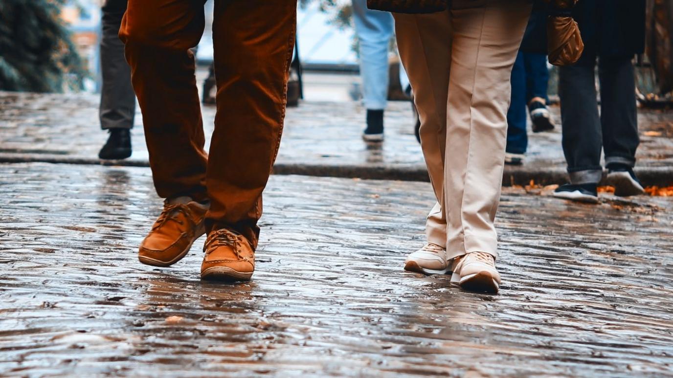 A close-up on the legs and feet of two people walking. On the left is someone wearing burgundy trousers and brown shoes. On the left is someone wearing cream-coloured trousers and light-coloured shoes. They are walking on a cobbled street which is wet from rain. In the background re the legs of other pedestrians. 