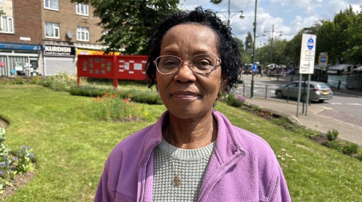 Norma Thompson in Wednesfield town centre. There are some shops and a car behind her. She has black hair and is wearing a jumper and purple fleece. She has a necklace on and is wearing glasses.