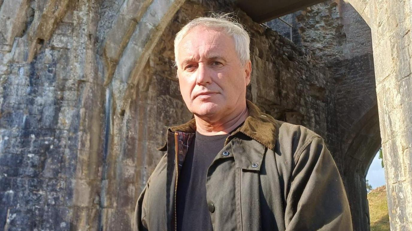 Rob Taylor, is Wales Wildlife and Rural Crime Co-ordinator, he wears a wax jacket and a black T-shirt next to the entrance of St Quentin's castle near Cowbridge. He has short grey hair and looks at the camera.